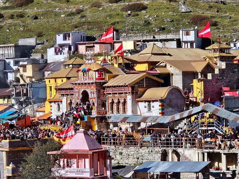 Badrinath Temple in bright sun: the painted Singh Dwar gate with its golden roofs and red-and-white flags, a dense crowd of pilgrims filling the forecourt and the railed queue below it, and the bare brown-green mountainside rising behind the town