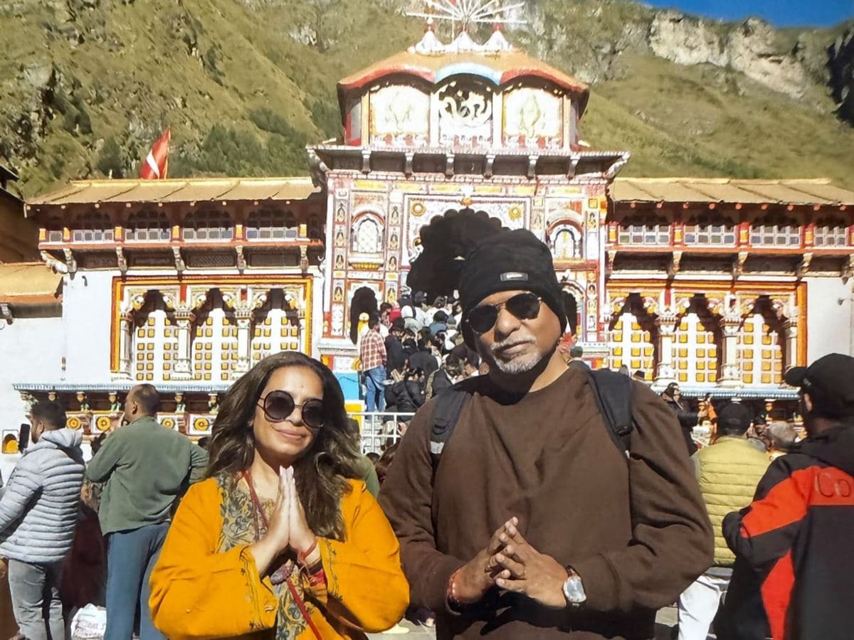 Sunirmal Ji, in a brown sweater and black woollen cap, and his wife, in a mustard-yellow kurta, stand with folded hands in front of the painted Singh Dwar of Badrinath Temple, pilgrims climbing the steps behind them under a clear blue sky