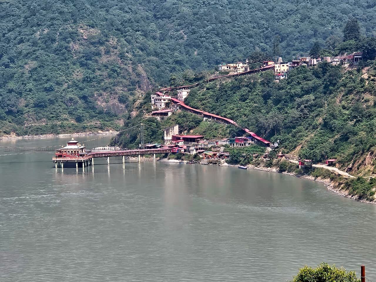 Dhari Devi Temple seen from across the Alaknanda: the shrine stands on pillars in the green-grey river, joined to the bank by a long bridge, and a red-roofed covered stairway climbs the forested hillside from the riverbank to the buildings at Kalyasaur on the highway above