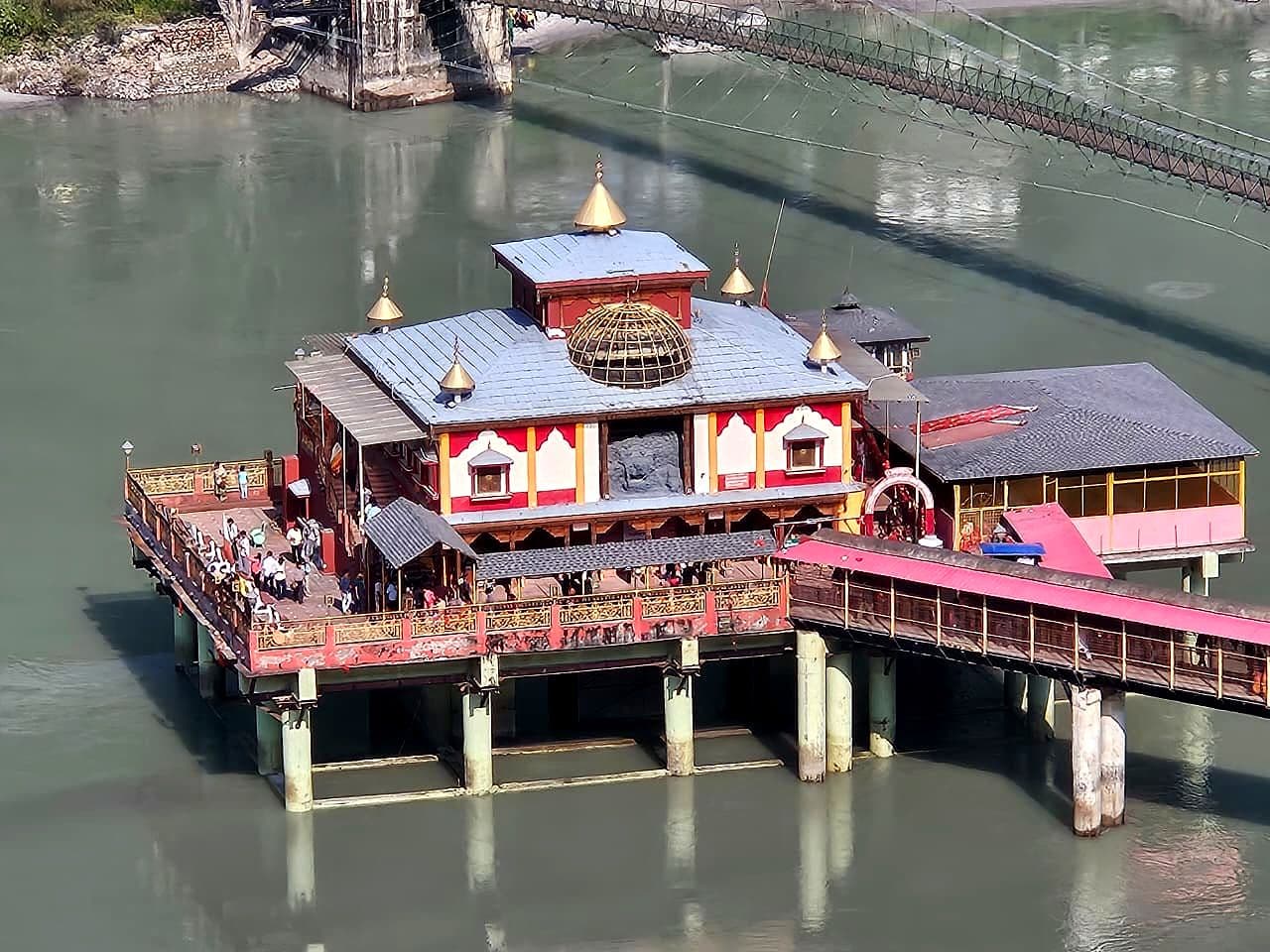 Close view of Dhari Devi Temple on its pillared platform in the Alaknanda: a red-and-white shrine with a grey roof, small golden finials and an open golden lattice dome, devotees on the railed terrace, and the pink-roofed covered bridge running in from the right