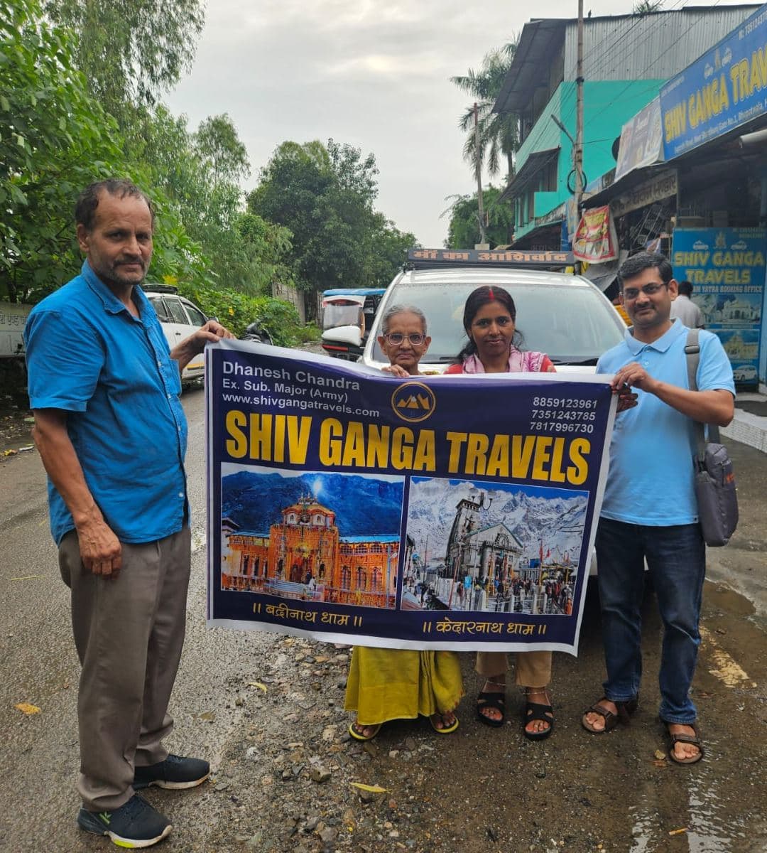 Dhanesh Chandra Mishra (left), founder of Shiv Ganga Travels, and a pilgrim family, an elderly mother, her son and his wife, hold a Shiv Ganga Travels banner showing Badrinath and Kedarnath temples beside a white car on a wet road outside the company office in Bhupatwala, Haridwar