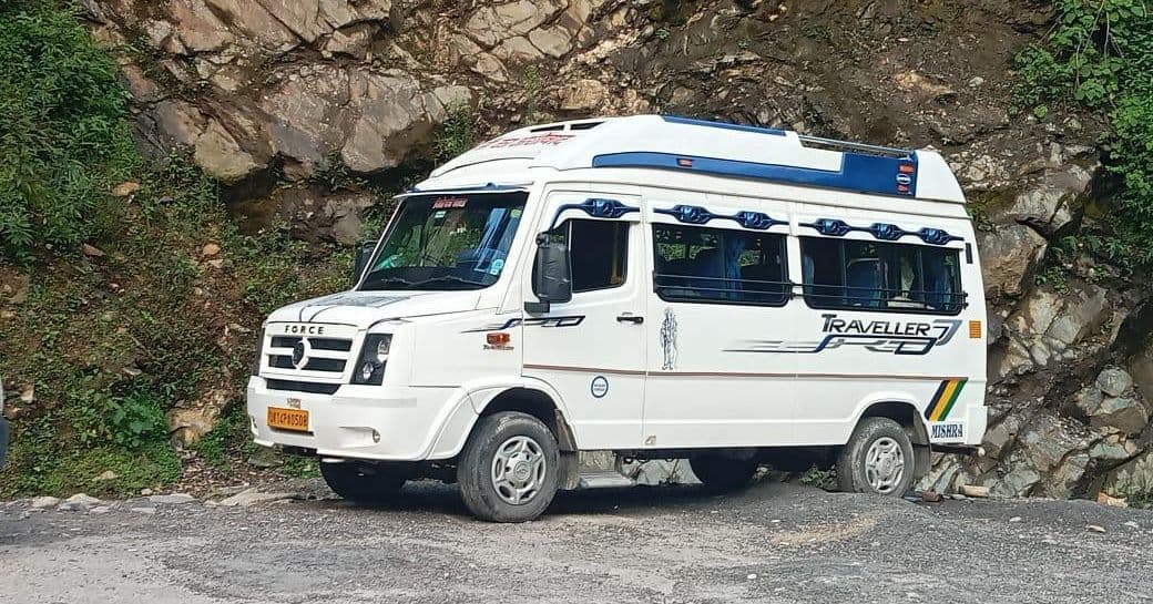 White Force Traveller high-roof van with a blue roof stripe and blue window trim, Traveller lettering on the side, a yellow commercial number plate and MISHRA lettered on the rear corner, parked on a gravel mountain road in front of a grey rock cutting with green slopes on either side